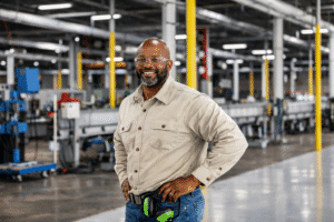 A man working. He is standing on the factory floor with his hands on his hips, smiling at the camera.